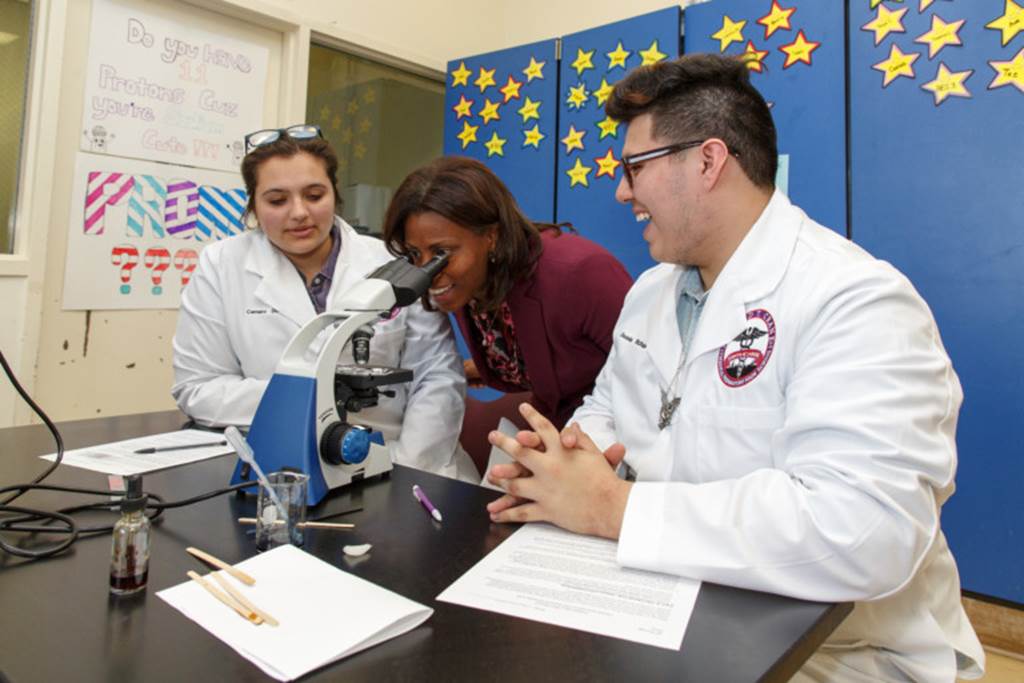 Two students observe as a woman looks into a microscope